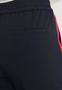 Pantalones de chándal azul marino con una cinturilla ajustada, que cuentan con un pequeño bolsillo trasero y una franja roja en el lado exterior de la pierna. Textura suave.