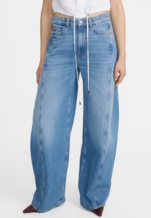 Frau trägt hellblaue Jeans mit weitem Bein, sichtbaren Nähten, Knopf, Reißverschluss und Taschen, kombiniert mit burgunderfarbenen spitzen Schuhen und einem bauchfreien weißen Top.