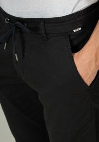 Schwarze Hose mit elastischem Bund und Kordelzug. Besitzt Seitentaschen, eine glatte Textur und ein kleines Markenetikett am Bund.