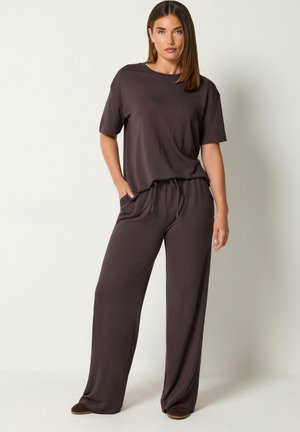 Femme debout portant un top ample à manches courtes marron foncé et un pantalon large assorti, les mains dans les poches, sur un fond uni.