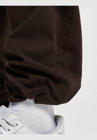Los pantalones marrones de puño presentan una tela suave y texturizada, combinados con calcetas blancas acanaladas y unas zapatillas blancas con suela texturizada.