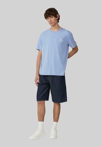 Lichtblauwe katoenen T-shirt met een ronde hals en korte mouwen, gecombineerd met donkerdenim korte broek. Witte sneakers en kousen van crew-lengte zichtbaar.