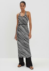 Schwarz-weiß gestreiftes, ärmelloses Kleid mit gebundenem Taillengürtel, fließendem Stoff und knöchellangem Design, kombiniert mit schwarzen weit geschnittenen Hosen und Sandalen.