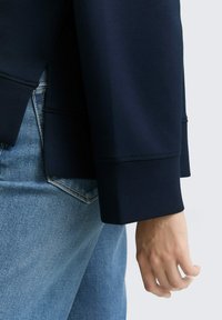 Manche de veste bleu marine avec une manchette structurée, associée à un denim bleu clair. Texture lisse, détails de couture minimaux au niveau de la manchette.