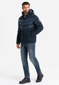 Marineblaue Steppjacke mit Chevron-Design, Kapuze und mit Reißverschlüssen versehenen Taschen. Weiches Material mit glatter Textur und robusten Hardware-Akzenten.