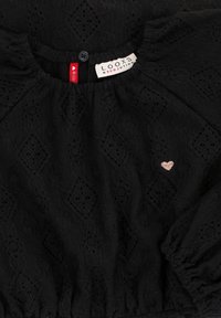 Zwarte textuurstof met diamantpatronen, elastische gathered halslijn en manchetten, klein rozenkleurig geborduurd hart, en twee zichtbare kledinglabels.