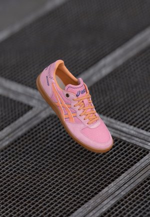 Pink-orangefarbener Asics-Sneaker mit Schnürsenkeln, liegend auf einer dunklen Metallgitteroberfläche.