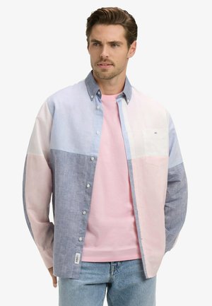 Homme portant une chemise à boutons à blocs de couleurs pastel sur un t-shirt rose et un jean bleu clair, regardant légèrement vers la droite sur un fond uni.