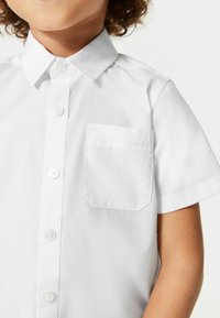 Chemise blanche à manches courtes avec un col structuré, fermeture à boutons sur le devant et une poche plaquée avec un logo en relief sur la poche.