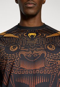 Μαύρο αθλητικό μπλουζάκι με περίπλοκο πορτοκαλί σχέδιο φυλετικού μοτίβου. Η γιακάς είναι μαύρος και ένα κίτρινο έμβλημα  βρίσκεται στο ύψος του λαιμού.