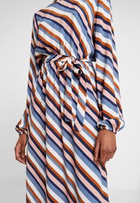 Robe rayée à manches longues, taille ceinturée, avec un mélange de couleurs bleu, rose, blanc et orange en motifs diagonaux. Tissu texturé.