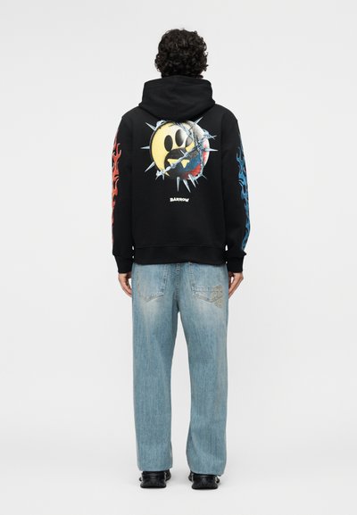 Persoană cu păr creț, purtând un hanorac negru cu un mesaj zâmbitor cu vârfuri, crăpat, și modele de flăcări roșii și albastre pe mâneci, blugi deschiși la culoare.