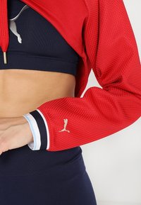 Rode sportieve jas met structuur, met een marineblauw crop top eronder. Heeft een gouden logo op de mouw en contrasterende blauwe en marineblauwe strepen op de manchetten.
