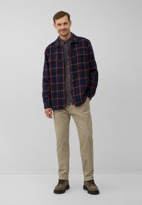 Glückliche Mann in einem navyblauen und roten karierten Jackett über einem gemusterten Hemd, beigen Hosen und braunen Schnürstiefeln, steht mit einer Hand in der Tasche.