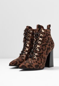 Paire de bottines à talons hauts imprimé léopard marron avec lacets noirs et œillets dorés sur une surface blanche contre un fond gris clair.