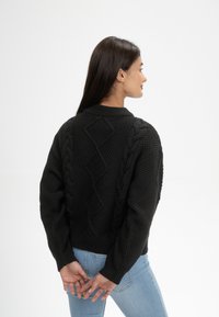 Schwarzer Strickpullover mit strukturiertem Muster und Zopfdetail auf dem Rücken, rundem Halsausschnitt und gerippten Bündchen. Kombiniert mit hellblauen Jeans.
