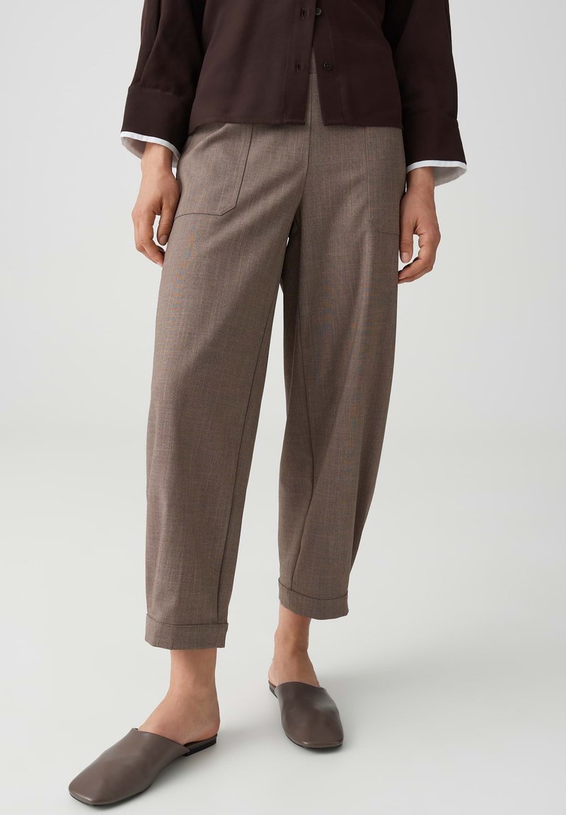 Woman wearing brown cropped wide-leg trousers with front pockets, paired with dark brown slip-on shoes and a dark top with white cuffs.
