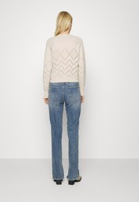 Suéter de punto color beige claro con cortes geométricos en la espalda, combinado con jeans rectos de mezclilla azul descolorido y botas negras de tacón cuadrado.