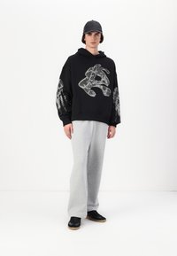 Svart hettejakke med grafisk design på forsiden og søleeffekter, kombinert med lysegrå joggebukser og svarte sneakers.