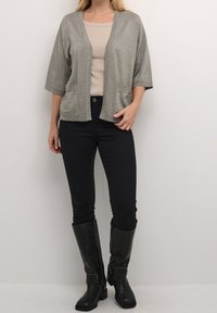 Zilveren metallic cardigan met open voorkant, korte mouwen en twee voorzakken, gecombineerd met een beige top en zwarte skinny jeans. Zwarte knielange laarzen.