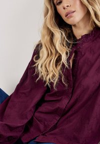 Blusa burdeos con un patrón floral texturizado, mangas largas, cuello alto y tapeta de botones, combinada con vaqueros.