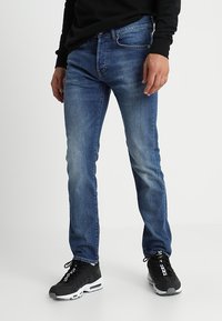 Niebieskie dżinsy o slim fit, z delikatnym wyblaknięciem, pięcioma kieszeniami i zamkiem błyskawicznym. Noszone z czarnymi trampkami.