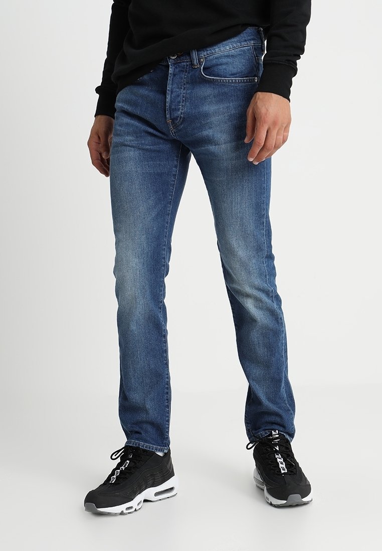 Niebieskie dżinsy o slim fit, z delikatnym wyblaknięciem, pięcioma kieszeniami i zamkiem błyskawicznym. Noszone z czarnymi trampkami.