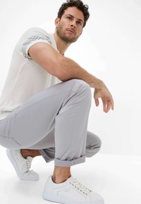 Pantaloni grigi chiari realizzati in tessuto liscio, abbinati a una camicia bianca a maniche corte con design a righe grigie e sneaker bianche.