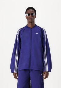 Veste de sport violette avec zip sur le devant, arborant des rayures blanches sur les manches et un petit logo blanc sur la poitrine. Tissu lisse et léger.