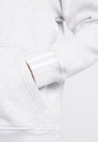 Hand i ljusgrå sweatshirtficka som visar ribbad manschett med två vita ränder på ärmen.