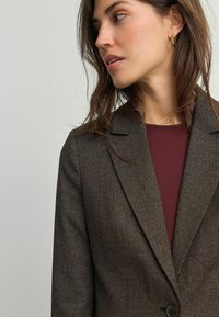 Brązowa marynarka w wzór houndstooth z klapami i zapięciem na jeden guzik, noszona na bordowej bluzce. Miękka tekstura i dopasowany krój.