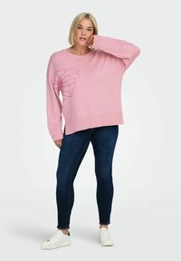 Maglione oversize rosa con scollo tondo, decorato con motivo floreale rosa chiaro, orlo a coste e maniche lunghe, abbinato a jeans scuri e sneaker bianche.