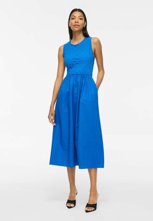 Vrouw draagt een mouwloze, felblauwe midi-jurk met een aansluitend, getextureerd lijfje en een geplooide rok, en staat met één hand in haar zak.