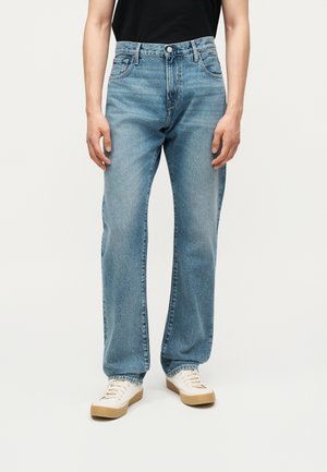 Osoba ubrana w jasnoniebieskie jeansy o prostych nogawkach i białe trampki, stojąca na jednolitym jasnym tle.