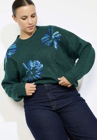 Maglione lavorato a maglia verde con design floreale in paillettes blu, collo rotondo e maniche lunghe, abbinato a jeans in denim scuro.