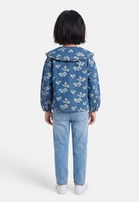 Enfant debout de dos portant une veste en jean bleue à fleurs avec un col volanté, un jean bleu clair et des baskets blanches sur un fond blanc.