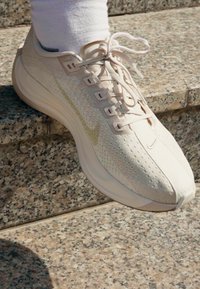 Hele beeži spordijalats, mis on valmistatud hingavast võrgust, millel on tekstuuriga pealsed, lame nöörid ja pehmendatud tald, asetatud kivistele treppidele.