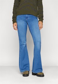 Jeans in denim blu flared con vita media, caratterizzati da una texture liscia e design classico a cinque tasche. Abbinati a scarpe verdi.