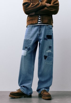 Plave traperice s opuštenim krojem, ukrašene komadićima tkanine u različitim nijansama plave boje i resama na rubovima. Nošene s smeđom obućom.