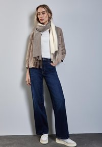 Beige Wildlederjacke, weißes Hemd, weite dunkelblaue Jeans, gemustertes grau-creamfarbiges Schal mitFransen und weiße Sneakers.
