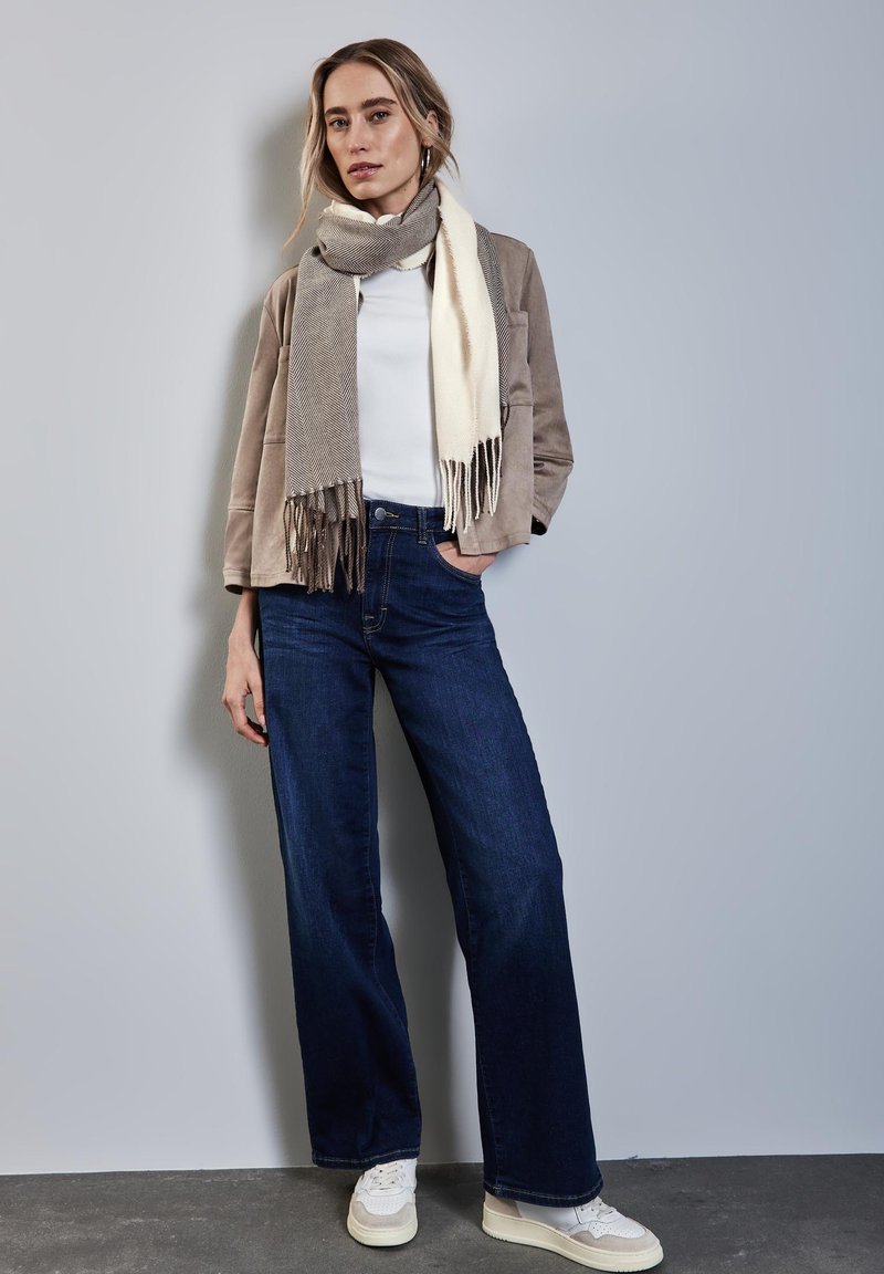 Beige Wildlederjacke, weißes Hemd, weite dunkelblaue Jeans, gemustertes grau-creamfarbiges Schal mitFransen und weiße Sneakers.