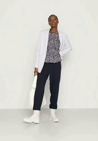 Blazer branco, top com padrão floral, calças azul-marinho e botas brancas; modelo segura também uma mala branca; fundo neutro.