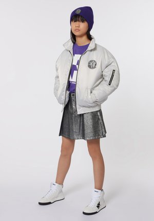 Zilveren bomberjack met logo, paarse beanie, grafisch paarse T-shirt, metallic zilveren rok en witte high-top sneakers.