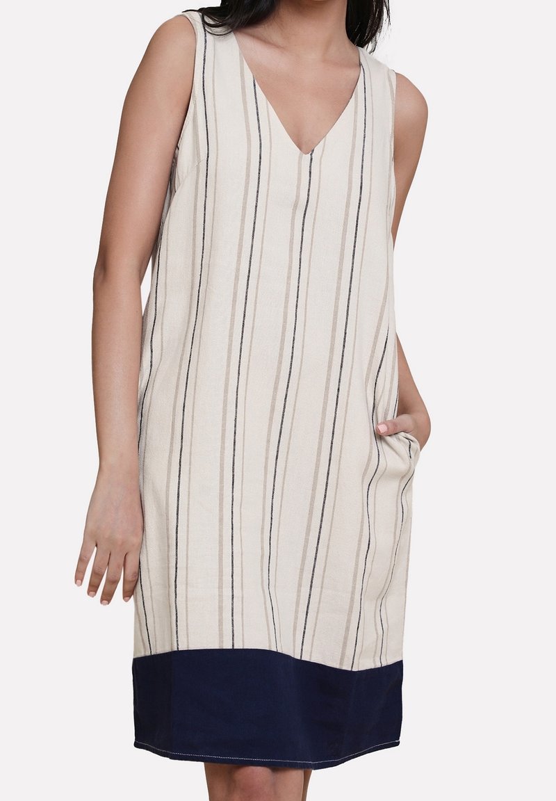 Robe beige sans manches avec des rayures noires verticales et un ourlet uni bleu foncé, portée par une personne avec une main dans la poche.