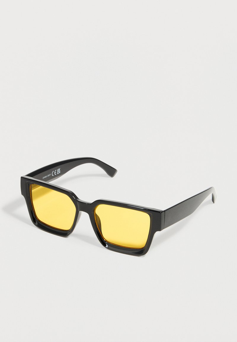 Lunettes de soleil à monture rectangulaire noire avec verres teintés jaunes reposant sur une surface blanche.