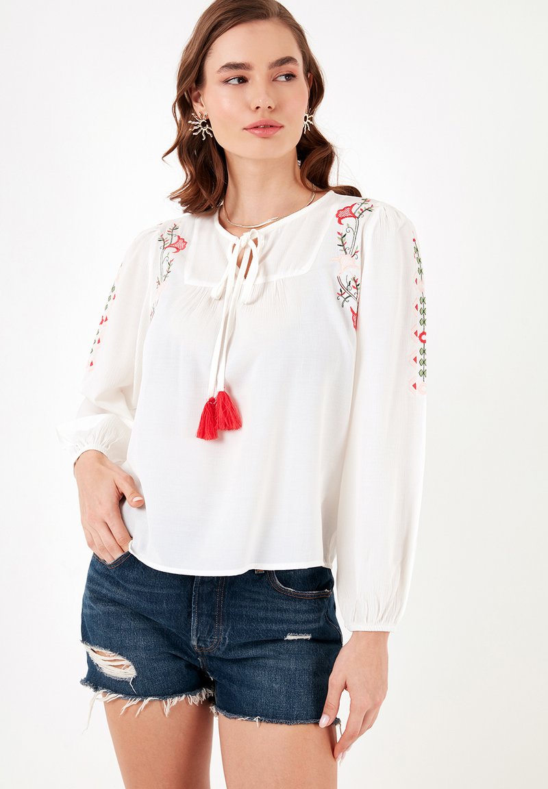 Weiße Bluse mit floraler Stickerei in Rot und Grün, gebundenem Kragen mit Quasten und langen Puffärmeln. Getragen mit zerrissenen Denim-Shorts.