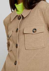 Karmelowa kurtka na guziki wykonana z miękkiego materiału, z kołnierzem, dwoma naszywanymi kieszeniami oraz dużymi brązowymi guzikami. Pod spodem widoczny jest neonowo zielony golf.