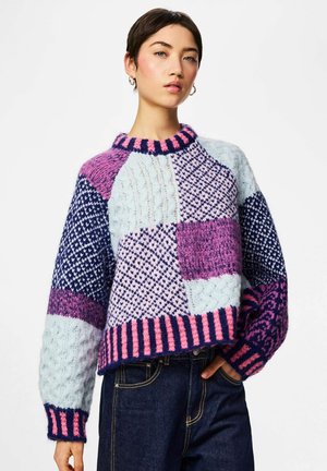 Frau trägt einen bunten Strickpullover mit Patchwork-Muster in Pink, Lila und Blau, kombiniert mit dunkelblauen Jeans, steht mit einer Hand in der Tasche.