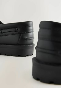 Chaussures en cuir noir avec un bout arrondi, un dessus texturé avec des détails de couture, et une semelle en caoutchouc épaisse et robuste avec des rainures.