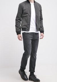 Šedá bomber bunda s černým žebrovaným límcem, v kombinaci s tmavě šedými džíny a černými koženými botami. Pod tím bílý tričko bez viditelných vzorů.